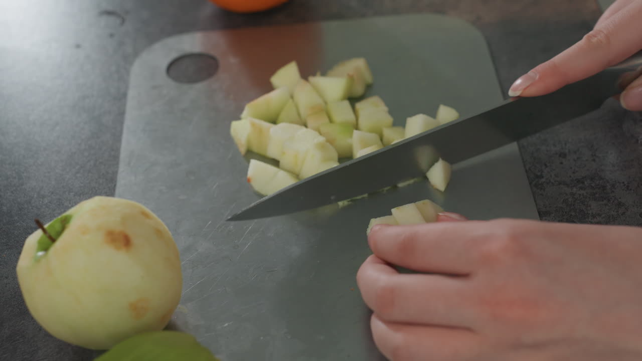 Mujer embarazada cortando una manzana en cubos sobre una tabla, vista aérea de las manos cortando una manzana pelada en cubos uniformes con un cuchillo, frutas naranjas y verdes cerca, encimera ordenada, preparación práctica y saludable para la dieta prenatal