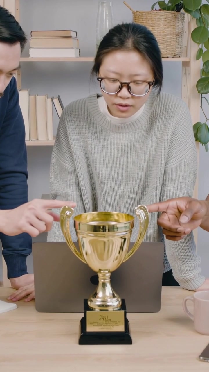 Vertical video: Left placing trophy urging woman grey sweater inspecting it at desk cheering by PC