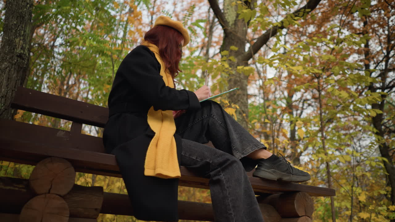 평화로운 자연 환경에 앉아있는 세련된 젊은 여자의 낮은 각도 뷰, 황색 베레트는 조용한 숲 환경에서 활기찬 가을 잎으로 둘러싸인 나무 벤치에 그녀의 그림에 초점을 맞추고 있습니다.