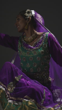 vertical video of female kathak dancer performing dance wearing traditional indian dress seated on floor in spotlight 3