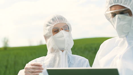 vista de cerca de investigadores caucásicos con traje protector sosteniendo un tubo de ensayo y usando una laptop mientras controlan plagas en el campo verde