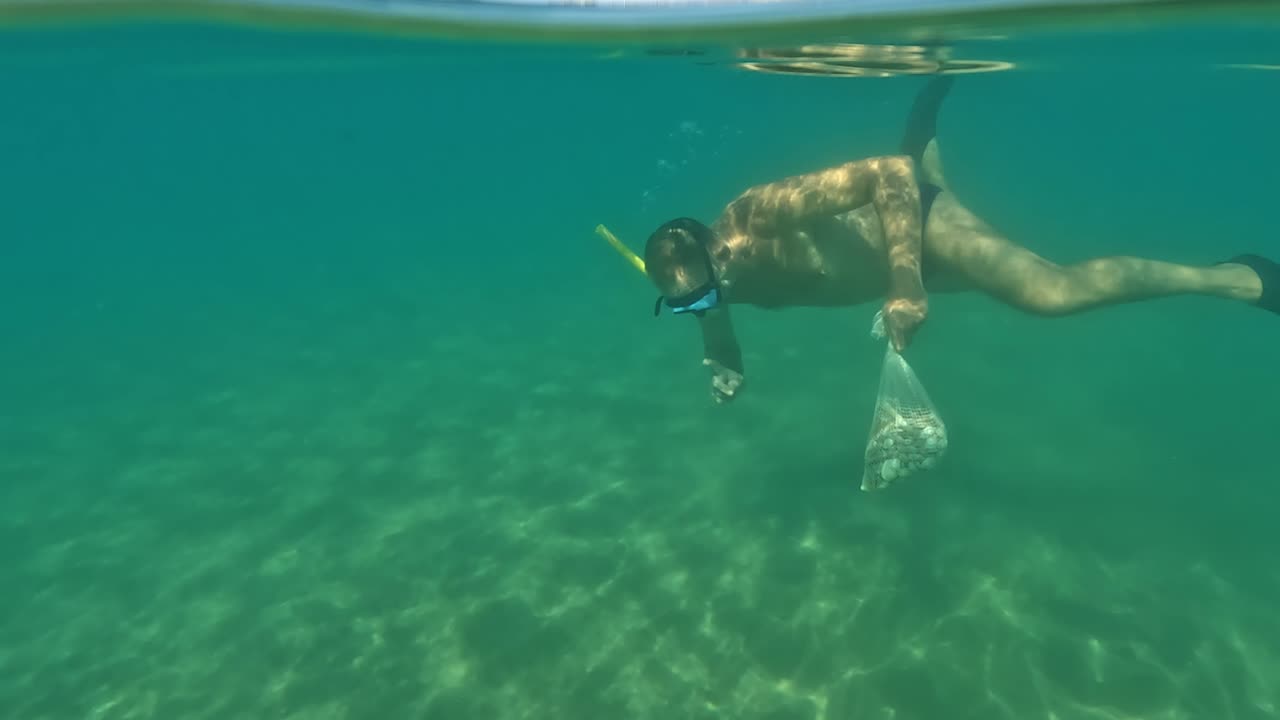 마스크와 스노클링 해산물로 잠수하는 어부의 절반 수중 전망, 이탈리아 배경에 트라보치 나무 플랫폼이 있는 결정질 바닷물에 조개로 가득 찬 가방을 들고