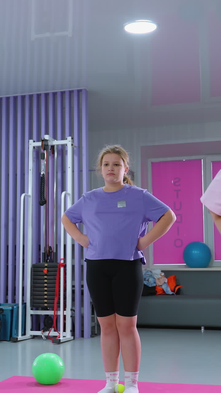 vista parcial de un instructor de fitness dirigiendo a estudiantes jóvenes en ejercicios de construcción de resistencia, manos sobre su cintura, en un gimnasio con esteras y equipos rosados en el fondo