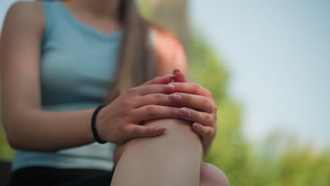 vista parcial de una mujer joven sentada al aire libre con las piernas cruzadas, las manos juntas en la rodilla, haciendo movimientos suaves con los dedos, usando un brazalete de mano negro, usando una camiseta cian, bajo la suave luz del sol
