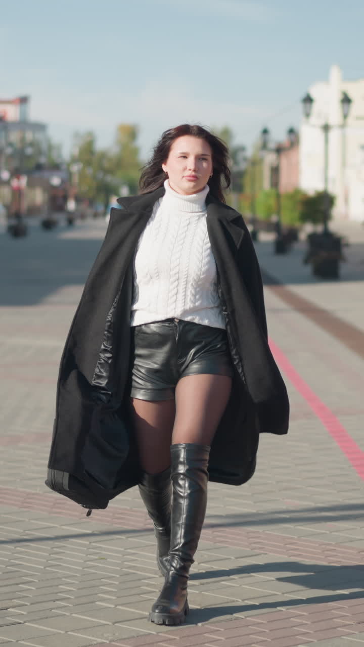 mujer confiada paseando en abrigo negro, suéter blanco y botas negras en una calle urbana pavimentada durante un día soleado, sosteniendo un bolso negro, rodeada de edificios modernos