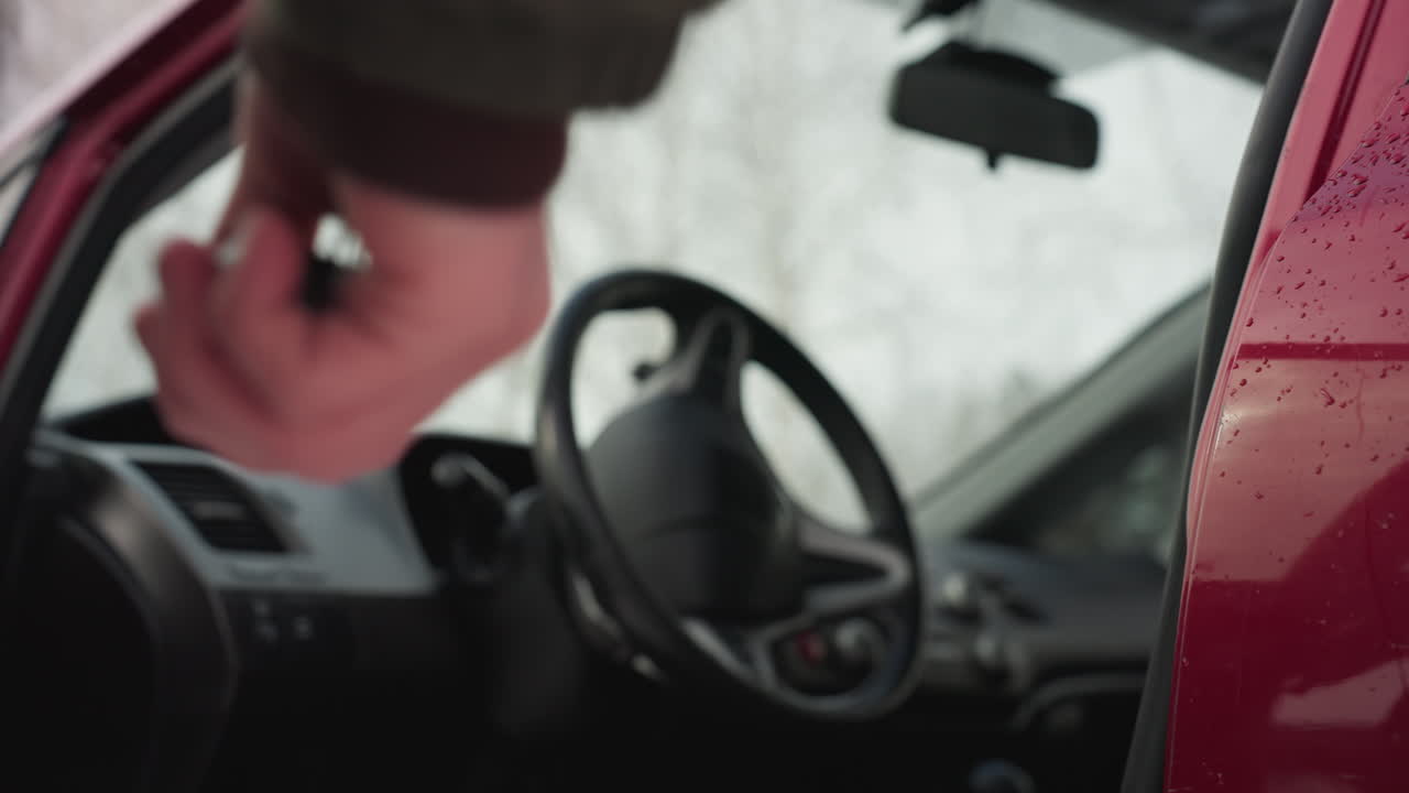 Close up of driver hand pulling chrome handle on red car door while preparing to enter vehicle, with water droplets on door surface and snowy background reflecting cold winter condition