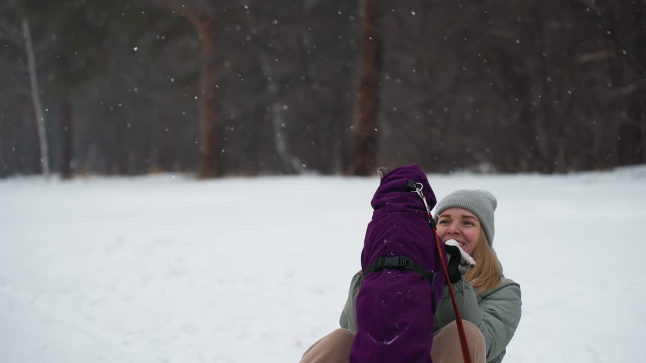 Woman in gray winter jacket and beanie smiles brightly while holding excited dog wearing purple coat in snowy park, enjoying playful interaction and expressing genuine happiness in cold weather outdoor setting