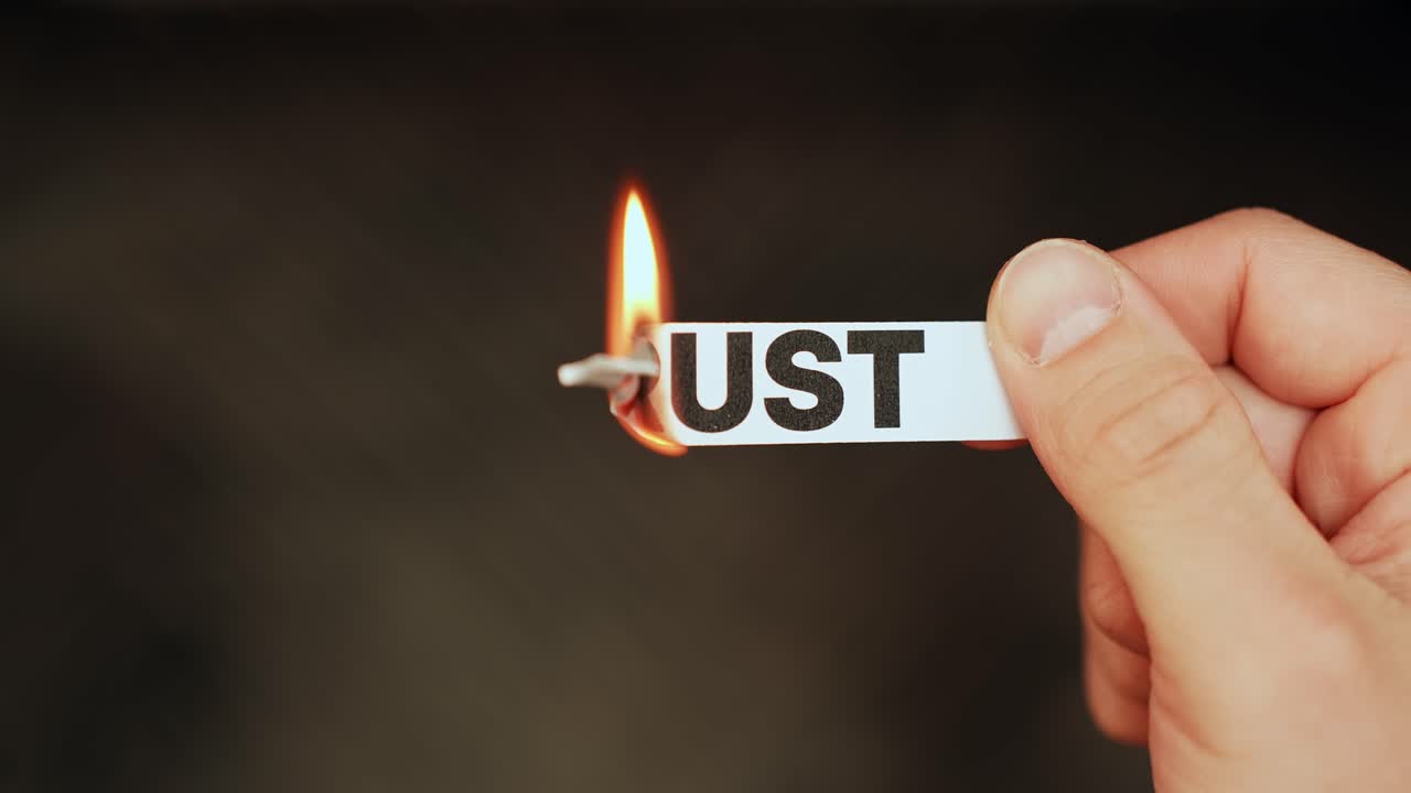 Close-up of a hand holding a paper strip labeled “TRUST” as the edge burns with flame on a dark background. Broken trust and relationship crisis concept with copy space