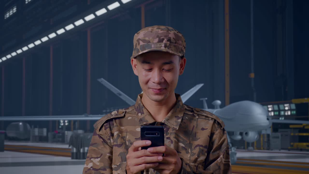 Primo piano di soldato asiatico che usa lo smartphone mentre si trova in piedi con un moderno drone militare in un hangar di manutenzione.