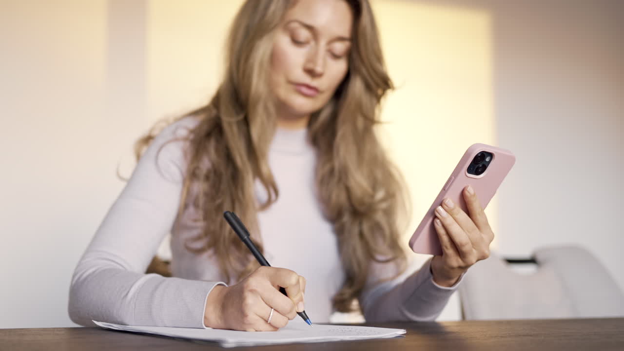 Woman writing on a piece of paper on a table while holding a phone