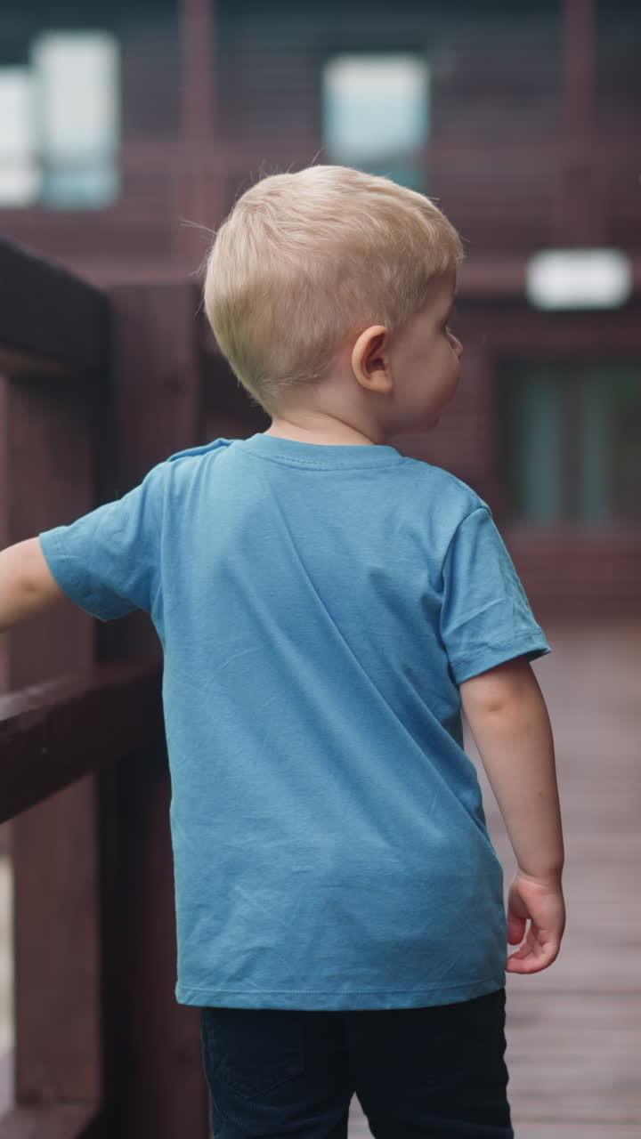 Sweet little boy walks holding handrail along wet veranda deck at wooden cottage house after warm spring rain backside view slow motion