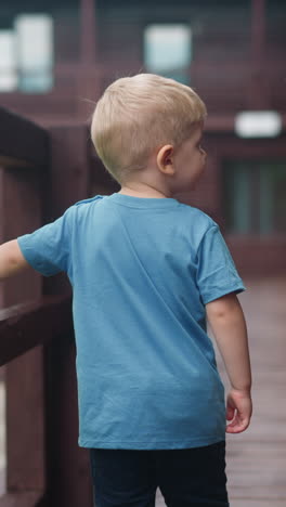 sweet little boy walks holding handrail along wet veranda deck at wooden cottage house after warm spring rain backside view slow motion