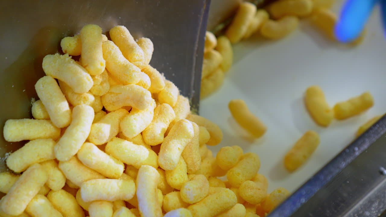 Light yellow crispy snacks falling on the conveyor belt. Corn sticks coated with sugar frosting close up.