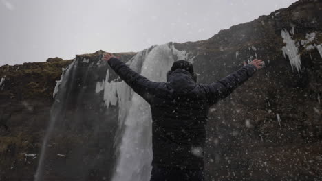Back-View-of-Man-in-Snowfall-Walking-Under-Waterfall-on-Cold-Winter-Day-and-Raising-Arms,-Slow-Motion