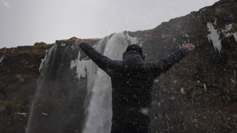 back view of man in snowfall walking under waterfall on cold winter day and raising arms, slow motion