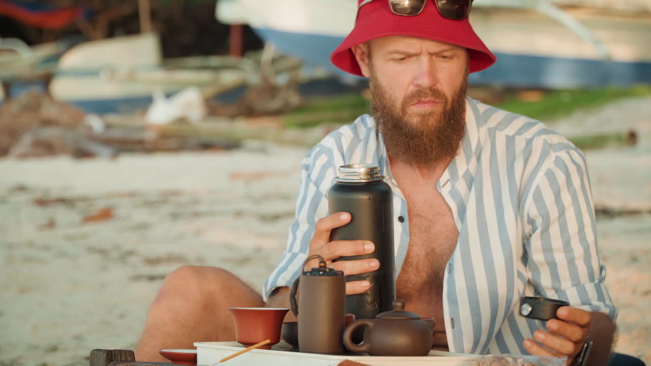 Rituale del tè solitario in spiaggia, uomo caucasico prepara una bevanda consapevole sulla riva, cappello da pescatore rosso e camicia a righe, thermos e teiera su un tavolo di legno, versamento costante, vapore che sale, pratica concentrata, all'aperto.