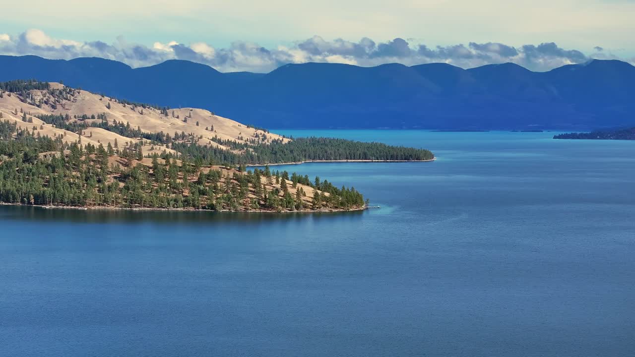 Costa arborizada e encostas de montanha acidentadas com vista para a água azul-profundo do lago sob um céu claro, criando uma paisagem alpina dramática, Flathead Lake, Montana, EUA.