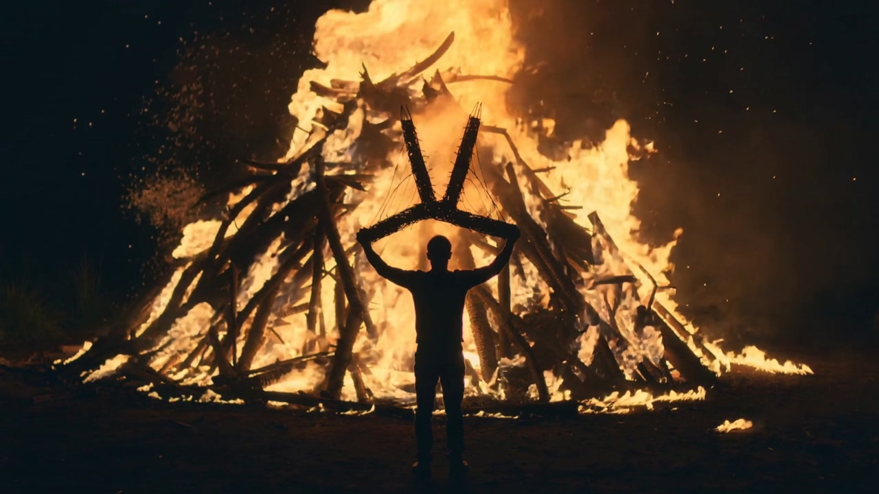 Raising lone man in dark clothing holding V-shaped wood emblem in night field, bonfire burning