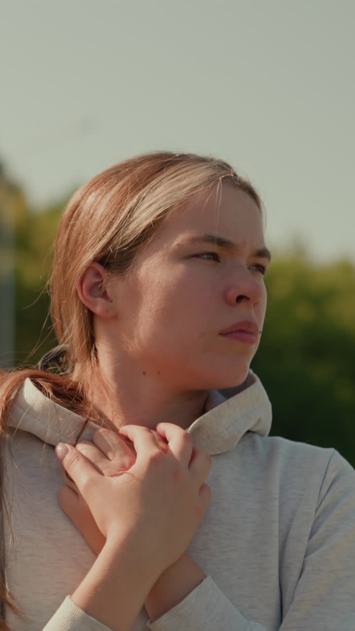 캐주얼한 후디를 입고 야외에 있는 사려 깊은 여인, 은 날 사람들, 나무, 기둥, 그리고 초록색의 흐릿한 배경으로 둘러싸인 빈 경기장 설정에서 사려 깊게 는 동안 후드를 붙잡고 있습니다.