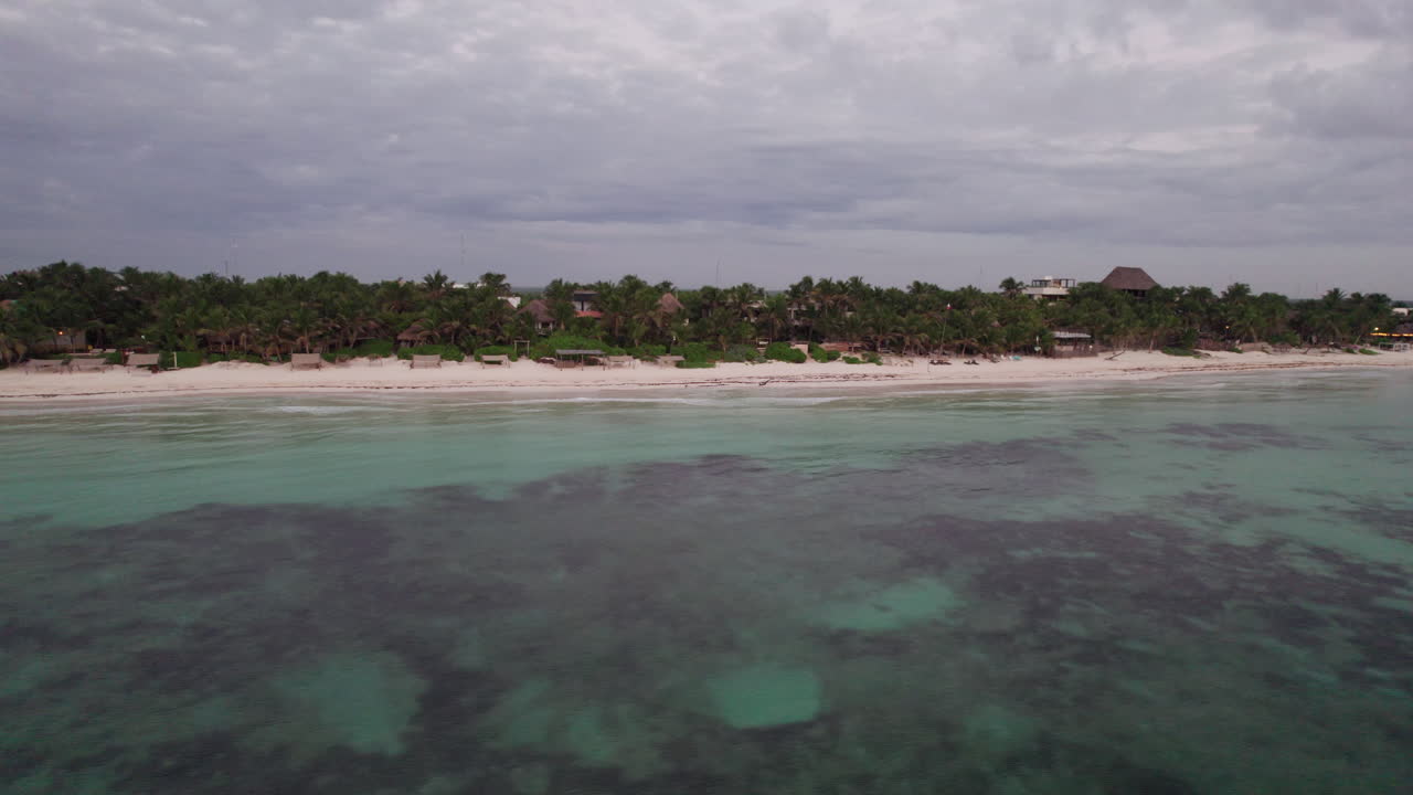 fotografía aérea de cabañas y chozas rodeadas de palmeras frente a una arena blanca y un océano azul cristalino en tulum, méxico