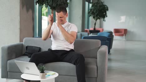 close-up-portrait-of-a-young-man-in-a-white-T-shirt-against-the-backdrop-of-a-business-center,-sitting-on-a-sofa,-passionately-following-a-sports-match-on-his-laptop