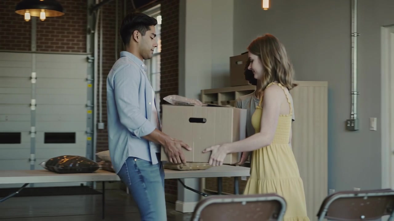 Reaching for box, blue-shirted man leading yellow-dressed woman lifting box across hall, stocking