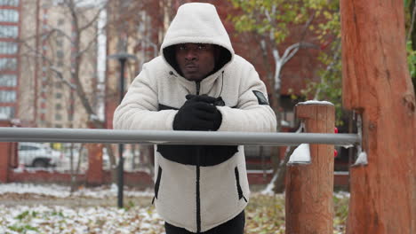 young man cracks knuckles in white hoodie outdoors, resting on iron bar with cold air visible from mouth, urban background with buildings, snowy ground, and wooden workout equipment