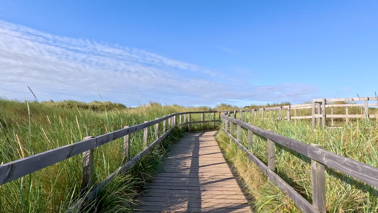 Smooth camera movement along sunlit boardwalk, surrounded by tall grass, blue sky, and sand dunes