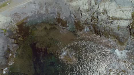 Top-down-aerial-of-tropical-ocean-water-waves-crashing-on-a-rocky-shoreline-during-a-summer-day