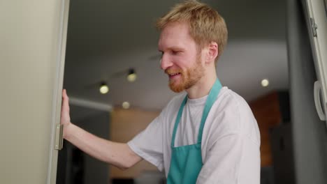 Close-up-a-blond-janitor-guy-in-a-white-T-shirt-and-blue-apron-washes-glass-walls-in-a-modern-apartment-during-cleaning