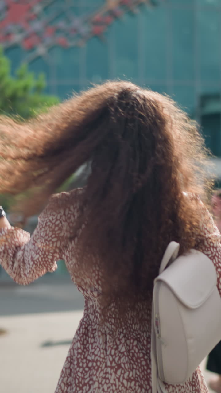 vista trasera de un adulto joven con cabello rizado, bolsa blanca, usando un brazalete de mano y un reloj inteligente, caminando en un área urbana con coches estacionados a ambos lados y personas en el fondo