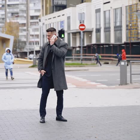 handsome man in coat talks the phone outdoors. young businessman standing in the street with a mobile phone and looking on handwatch. slow motion.