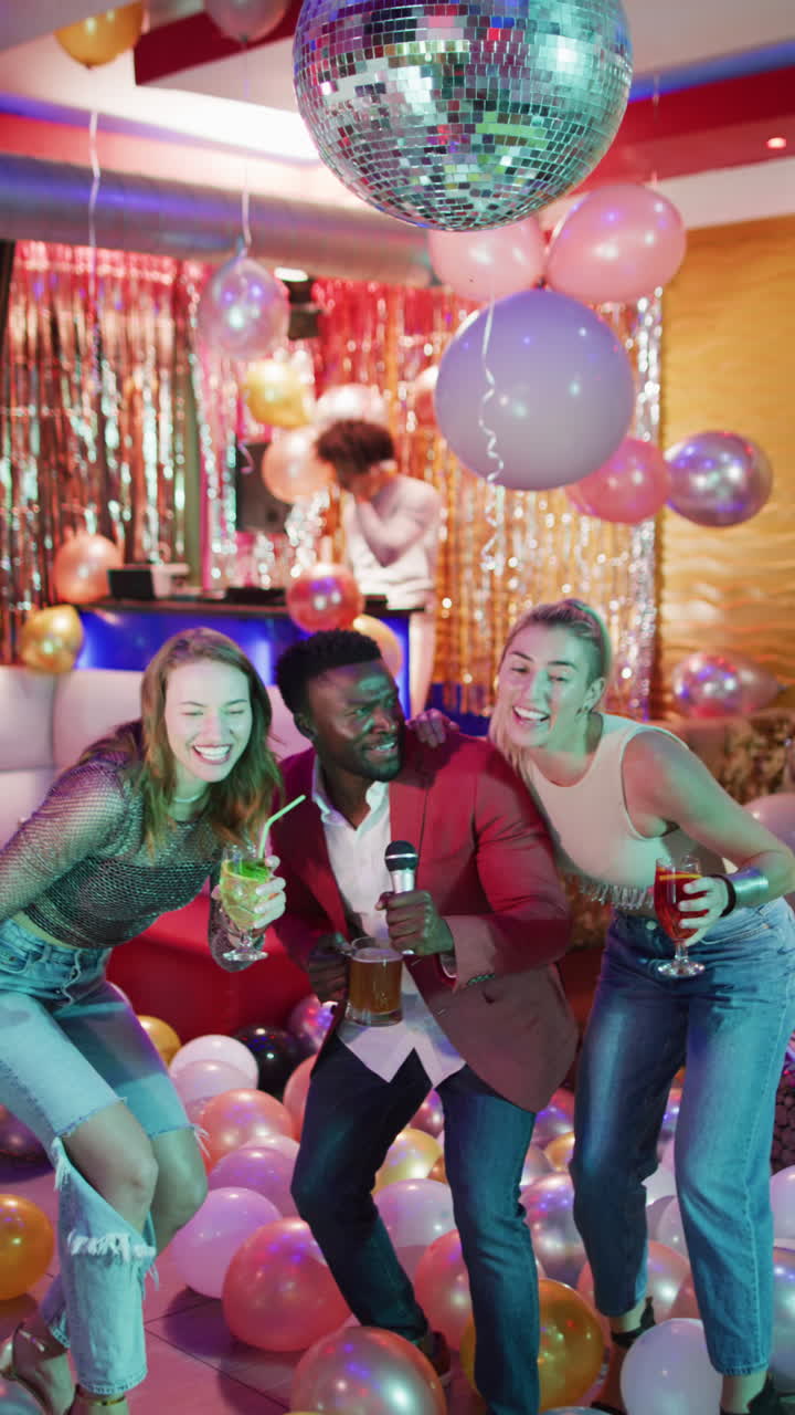 Vertical video of happy, diverse group of friends singing karaoke with drinks at a nightclub bar