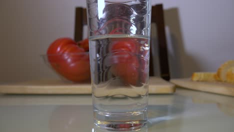 a glass of clean water on the table with big red tomatoes and bread in the background - closeup shot