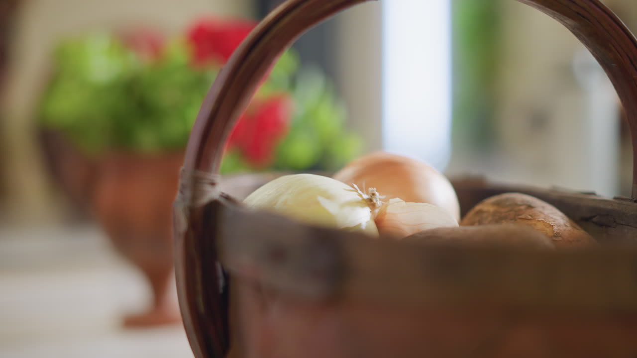 Basket of onions and potatoes on countertop, soft morning light, potted red geranium blurred in background, cozy home cook prep scene with rustic woven basket and fresh produce ready for recipe