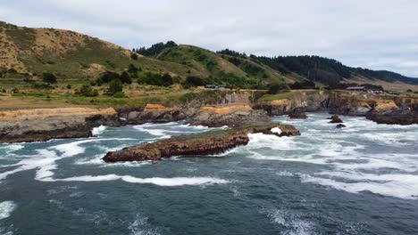 Oregon:-Rastreando-Una-Roca-Costera-Cubierta-De-Musgo-Mientras-Las-Olas-Del-Océano-Chocan-Contra-La-Roca-Oceánica-Y-Las-Aves-Gaviotas-Vuelan-Por-Los-Bosques-Pnw-Y-El-Fondo-épico-De-La-Granja,-Agua-Azul-Turquesa,-Lagunas-Marinas