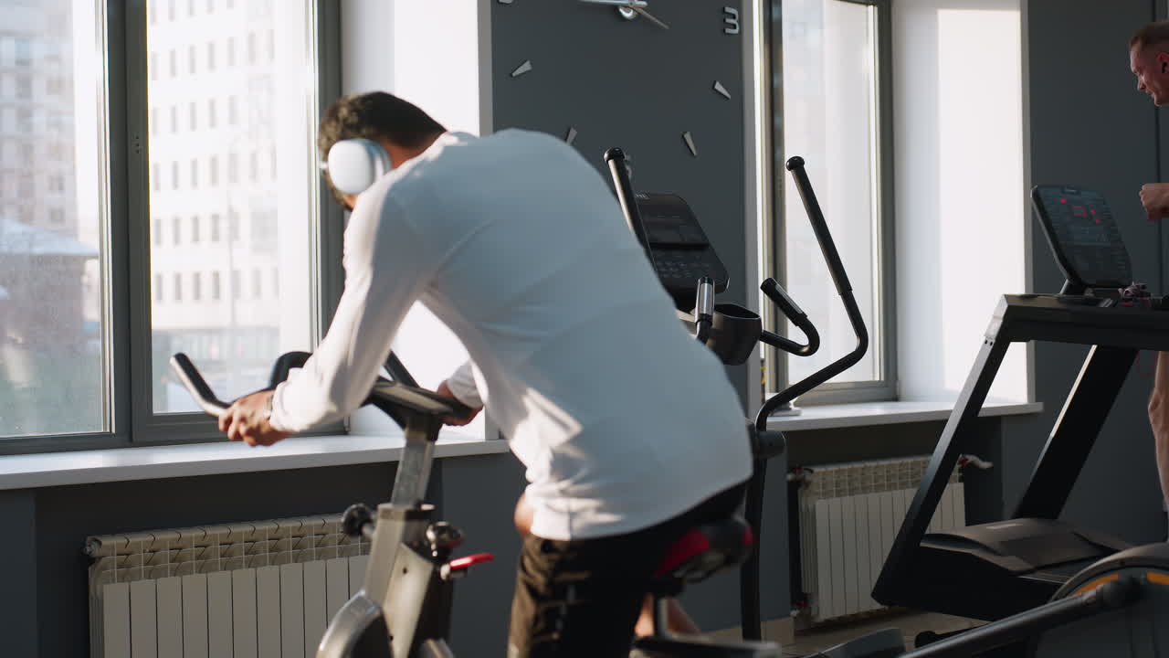 Young student in white sportswear walks to stationary bike and climbs on to begin cycling under sunlight streaming through gym windows, while background shows equipment and blurred workout scene