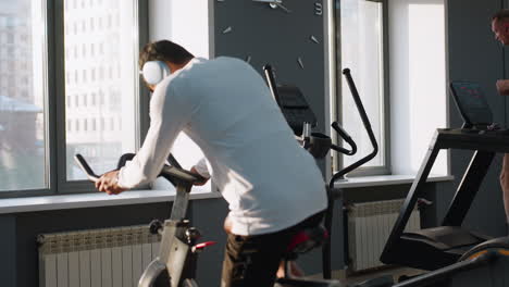 young student in white sportswear walks to stationary bike and climbs on to begin cycling under sunlight streaming through gym windows, while background shows equipment and blurred workout scene
