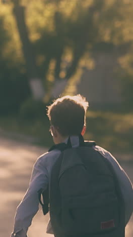 young pupil in grey shirt with large black bag runs along asphalt road and throws paper sheets slow motion backside view