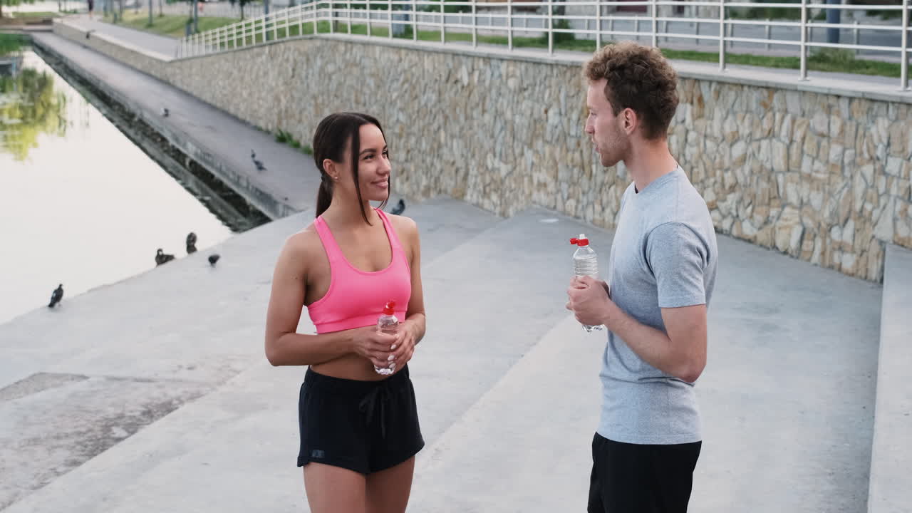 feliz pareja deportiva bebiendo agua y hablando entre ellos mientras toman un descanso durante su sesión de carrera en la ciudad
