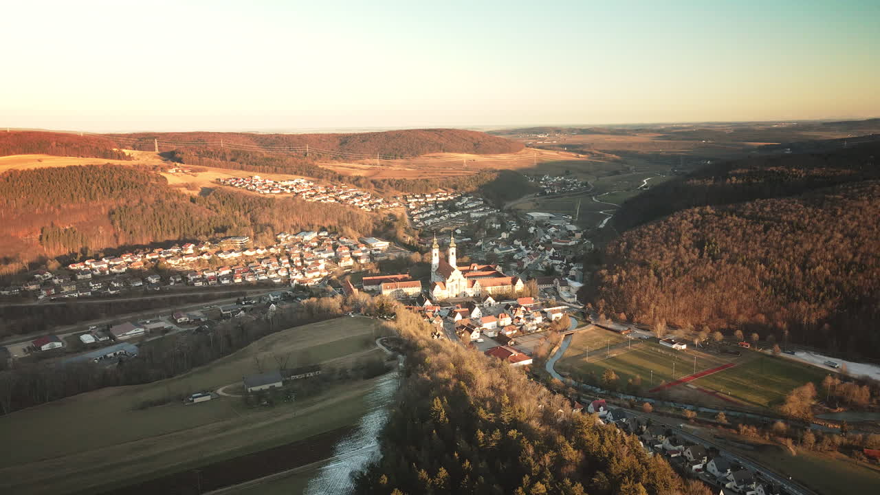 Drone advances forward over Zwiefalten's quaint rooftops toward hilltop monastery towers amid autumn hills, fields and river in warm Swabian Alb sunset. Timeless baroque panorama