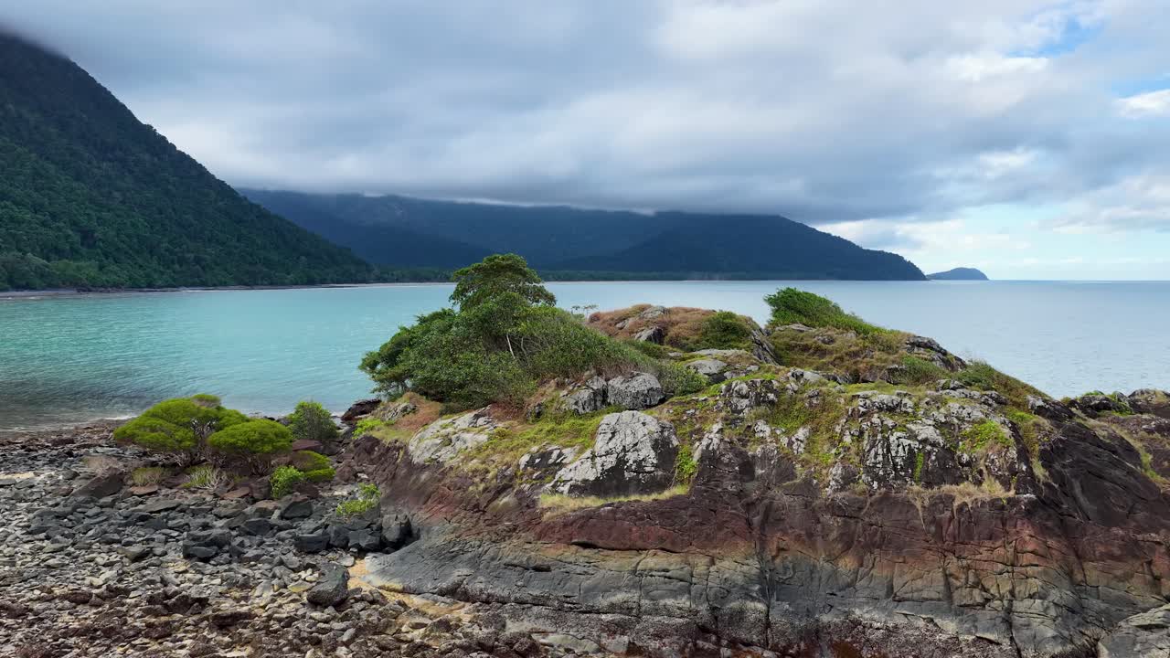 A tranquil island with lush greenery surrounded by turquoise waters and mountainous backdrop under cloudy skies