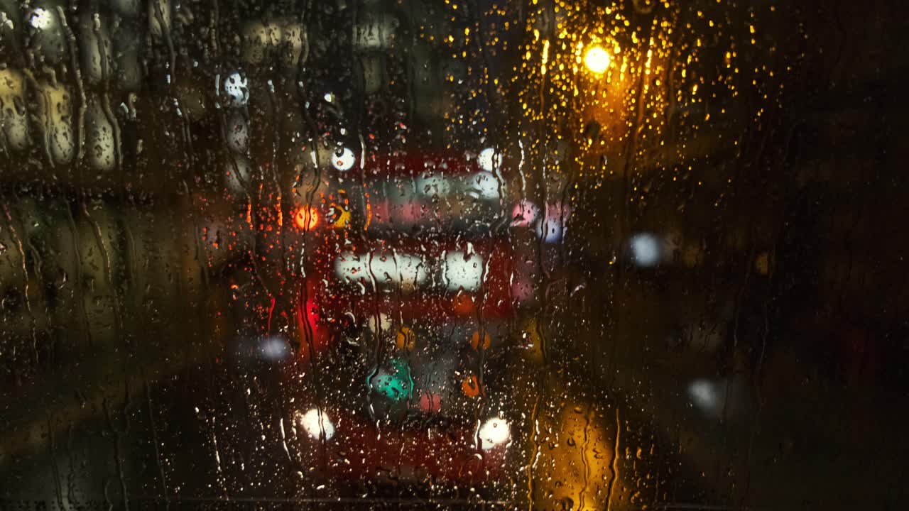 Rain falls down back window of London bus as buses and traffic pass behind out of focus