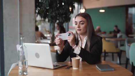 Una-Mujer-Alegre-Sentada-En-Un-Elegante-Café-Vestida-De-Negocios-Con-Una-Computadora-Portátil,-Jugando-Felizmente-Un-Juego-En-Su-Teléfono-Inteligente-Con-Una-Sonrisa