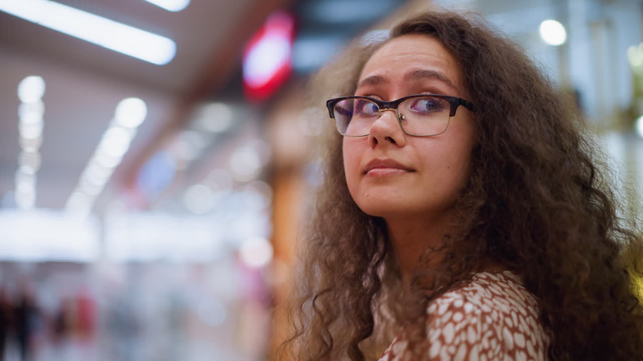mujer con gafas y cabello rizado mirando a su alrededor en un centro comercial brillantemente iluminado, rodeado de suaves luces bokeh, con detalles de fondo borrosos de las luces del centro comercial y la gente