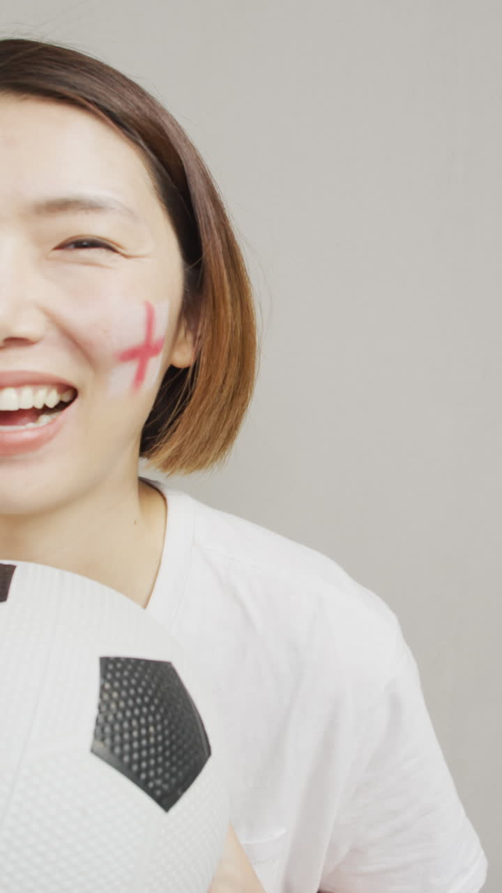 Vertical video of happy asian woman holding football with flag of england on cheek