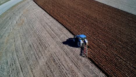 Toma-Aérea-Enfocada-En-Un-Tractor-Azul-Arando-Un-Campo-Cambiando-El-Color-Del-Suelo