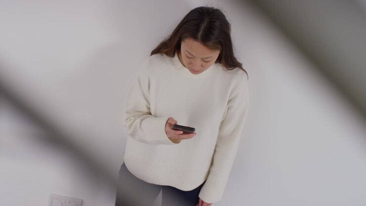 Ovenfra skud af stresset eller ængstelig kvinde, der læner sig op ad en væg derhjemme og reagerer på internet- eller sociale medier-nyheder, besked eller historie på mobiltelefon 1