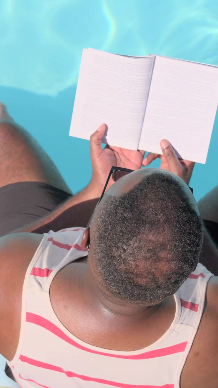 Vertical video: Adjusting man in striped tank holding open notebook to read at pool, sunglasses