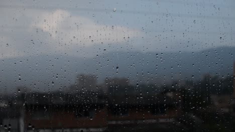 Regentropfen-Fallen-Stetig-Durch-Ein-Fensterglas-Mit-Der-Stadt-Im-Hintergrund